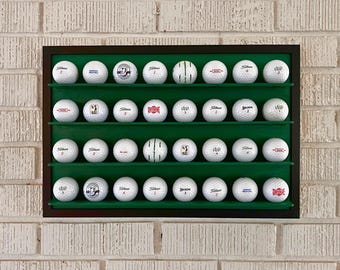 May include: A green display case with four shelves holds a collection of 24 white golf balls. The balls are arranged in rows and feature various logos and branding, including "Titleist", "Srixon", and "Top-Flite".