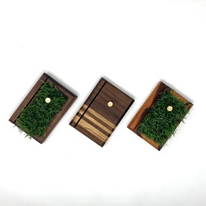 May include: Three rectangular wooden planters. Two have green artificial grass and a gold-coloured circular accent. The third has a striped design and a gold-coloured circular accent. The planters are made of dark brown wood.
