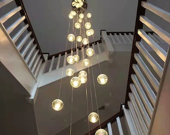 May include: A long, cascading chandelier with multiple clear glass globes hanging from a central point. The chandelier is suspended from the ceiling of a staircase.