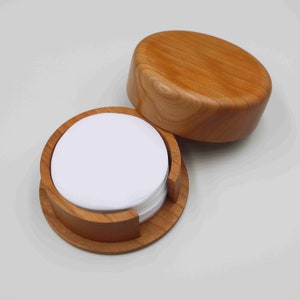 Può includere: Un set di sottobicchieri rotondi bianchi in un porta-bicchieri di legno con un coperchio. Il porta-bicchieri è in legno marrone chiaro con una finitura liscia.