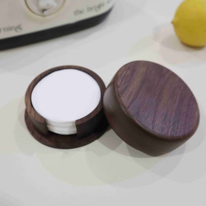 Puede incluir: Un juego de posavasos redondos de madera con un acabado marrón oscuro. Los posavasos están apilados y tienen un inserto de papel blanco.