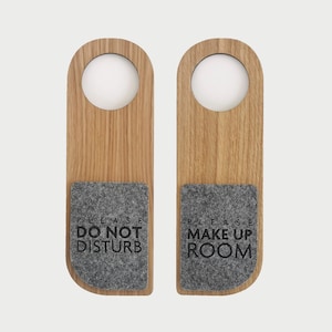 以下が含まれることがあります： 灰色のフェルトのインサートが付いた木製のドアハンガーが2つ。左側のハンガーには「邪魔しないでください」と書いてあり、右側のハンガーには「部屋を整えてください」と書いてあります。