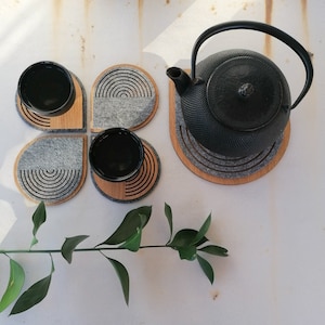 May include: Four gray felt coasters with a geometric pattern, each with a small, round, black ceramic dish. A black cast iron teapot sits on a wooden coaster with a similar geometric pattern.