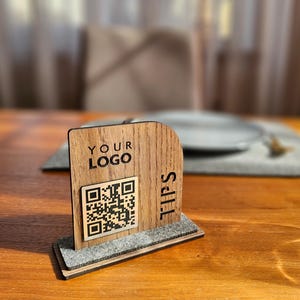Puede incluir: Soporte de mesa de madera con las palabras "YOUR LOGO" y "TIPS" impresas. Se muestra un código QR en la parte delantera. El soporte está sobre una base de fieltro gris, sobre una mesa de madera.