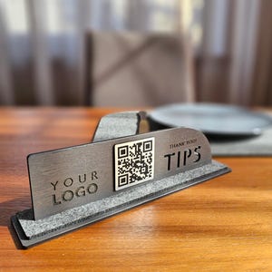 Könnte beinhalten: Ein dunkler Tischaufsteller aus Holz mit einem QR-Code und den Worten "YOUR LOGO" und "TIPS". Der Aufsteller steht auf einem grauen Filzsockel. Die Worte "THANK YOU!" stehen über dem Wort "TIPS".