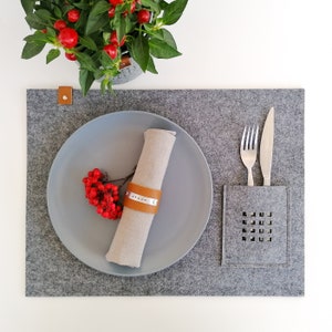 May include: A grey felt placemat with a grey plate, a linen napkin with a leather napkin ring, and a silver fork and knife in a grey felt pocket.