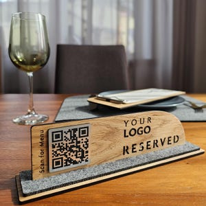 Könnte beinhalten: Holztischschild mit einem QR-Code und dem Text "Scan for Menu" und "YOUR LOGO RESERVED". Das Schild steht auf einer grauen Filzbasis. Ein Weinglas und eine Speisekarte befinden sich ebenfalls auf dem Tisch.