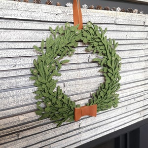 Puede incluir: Una corona de fieltro verde con un lazo de cuero marr&oacute;n colgada de una pared de ladrillo blanco. La corona est&aacute; hecha de hojas de fieltro y tiene un aspecto r&uacute;stico.