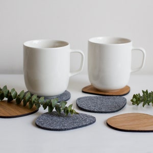 May include: Two white ceramic mugs on gray felt coasters with wooden coasters underneath. The mugs are on a white surface with green sprigs of eucalyptus.
