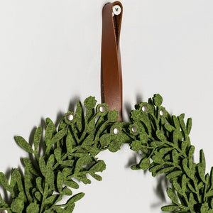Puede incluir: Una corona de fieltro verde con un lazo de cuero marr&oacute;n para colgar. La corona est&aacute; decorada con peque&ntilde;as hojas verdes y detalles met&aacute;licos plateados.