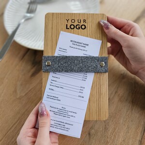 Könnte beinhalten: Ein hölzerner Rechnungsständer mit einem grauen Filmband und einer Quittung mit dem Text "RESTAURANT NAME THE PLAZA NAME" und dem Text "Vielen Dank für Ihren Besuch!" sowie einem Gesamtbetrag von 53,46 €.