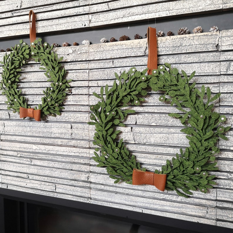 Puede incluir: Dos coronas de fieltro verde con lazos de cuero marr&oacute;n y lazos de fieltro marr&oacute;n colgando de una pared de ladrillo blanco.