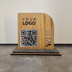Peut inclure: Panneau WiFi en bois avec un code QR et les mots "YOUR LOGO". Le panneau est en bois brun clair et comporte un code QR argenté. Le mot "WIFI" est découpé dans le bois. Le panneau repose sur une base en feutre grise.