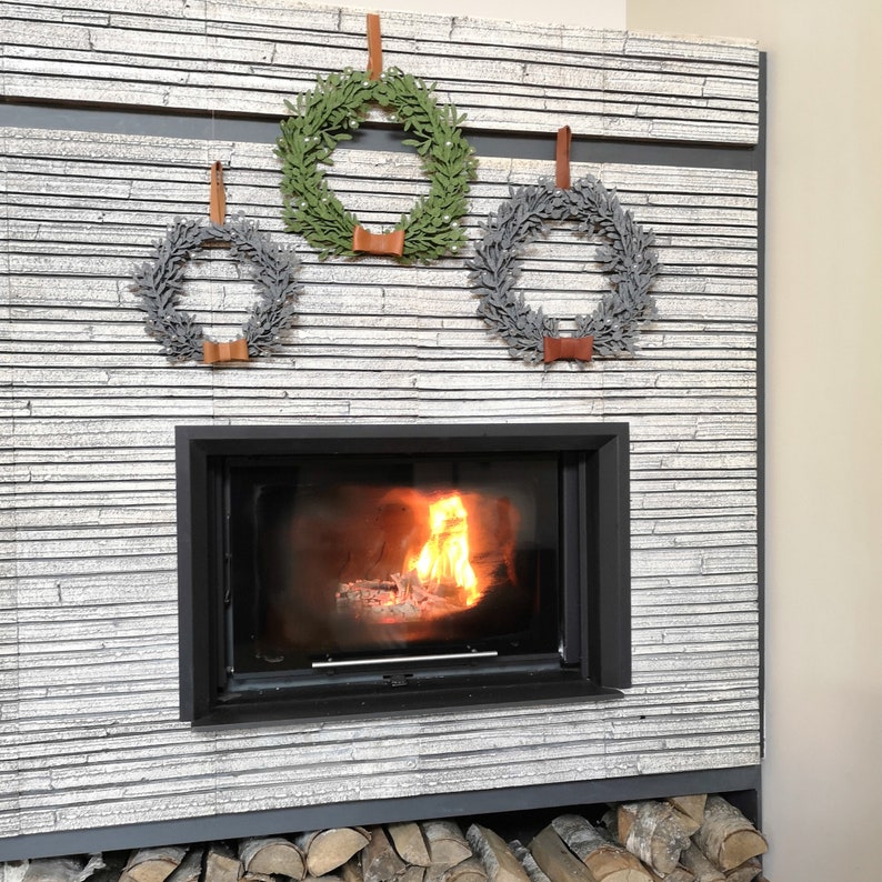 Puede incluir: Tres coronas, una verde y dos grises, cuelgan sobre una chimenea con un fuego crepitante. Las coronas est&aacute;n decoradas con bayas blancas y tienen lazos de cuero marr&oacute;n.