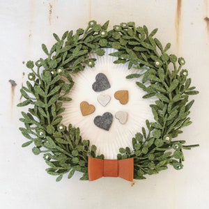 Puede incluir: Una corona de fieltro verde con un lazo marr&oacute;n y un fondo blanco. La corona est&aacute; decorada con peque&ntilde;os corazones de fieltro gris y marr&oacute;n.