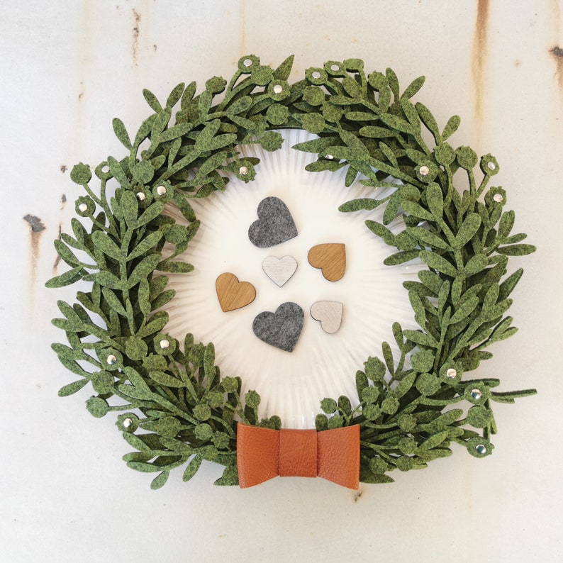 Puede incluir: Una corona de fieltro verde con un lazo marr&oacute;n y un fondo blanco. La corona est&aacute; decorada con peque&ntilde;os corazones de fieltro gris y marr&oacute;n.