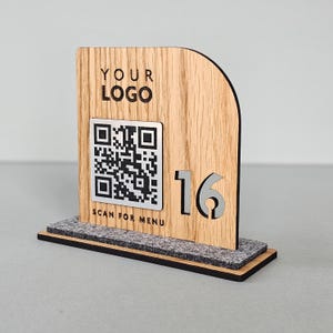 May include: Wooden table number with a QR code and the text "YOUR LOGO" and "SCAN FOR MENU". The number 16 is cut out of the wood. The base is a dark grey felt.