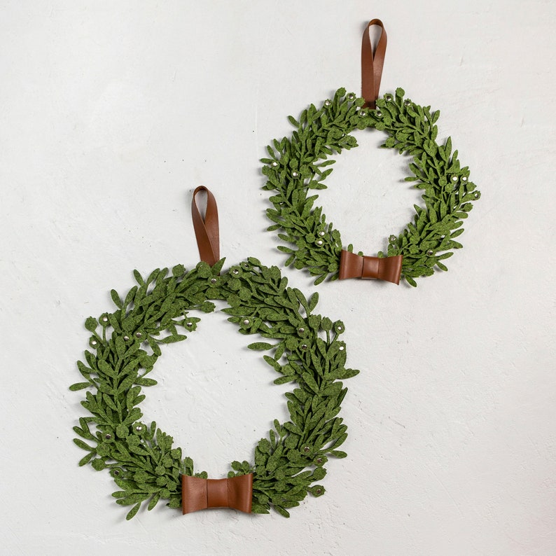 Puede incluir: Dos coronas de fieltro verde con lazos de cuero marr&oacute;n. Las coronas est&aacute;n decoradas con peque&ntilde;as cuentas plateadas.