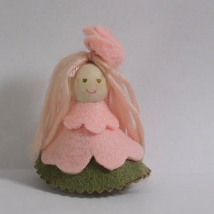 May include: A handmade doll with a wooden head, pink felt dress, and long pink yarn hair. The doll has a green felt base and a small pink flower on top of its head. The face is painted with a smiling expression.