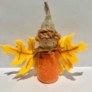 May include: A whimsical autumn-themed figurine. It features a wooden head with a smiling face, a hat made from an acorn cap, and orange felt clothing. The figurine has yellow leaf-shaped wings and a brown scarf.