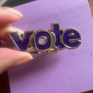 May include: A gold-colored metal pin with the word "VOTE" in purple enamel. The pin is held against a purple background. The pin is a political statement.