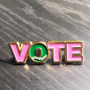 Op de afbeelding: Een goudkleurige pin met het woord "VOTE" in roze letters. De letter "O" is groen met een cirkel van kleine, heldere steentjes. De pin ligt op een donker, getextureerd oppervlak.
