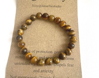 Puede incluir: Un brazalete marrón hecho de cuentas de ojo de tigre. El brazalete está en una tarjeta que dice "Tiger's Eye" y "Etsy@lunarfaestudio". La tarjeta también incluye texto sobre las propiedades protectoras del ojo de tigre.