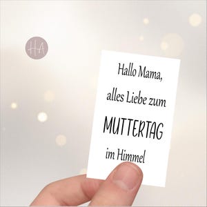 Op de afbeelding: Een witte kaart met de tekst "Hallo Mama, alles Liebe zum MUTTERTAG im Himmel" in zwart schrift. De kaart wordt door een hand vastgehouden tegen een zachte, vage achtergrond met bokeh-lichten. Een klein, cirkelvormig logo staat in de linkerbovenhoek.
