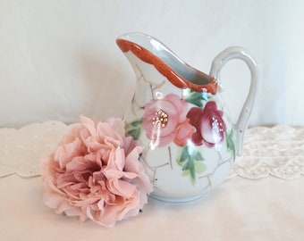 May include: A small white ceramic creamer with a floral design. The creamer has a handle and a spout. The flowers are pink, red, and green. The creamer is sitting on a white lace tablecloth.