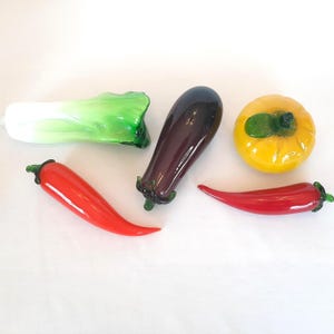 May include: A collection of glass vegetable sculptures, including a bok choy, eggplant, and a yellow squash. Two red chili peppers are also present. The glass vegetables are brightly colored and appear to be handmade.
