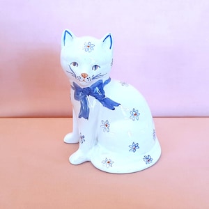 May include: A white ceramic cat figurine with blue accents. The cat has a blue bow around its neck and is decorated with small blue and red floral patterns. The cat is sitting upright with its tail curled.