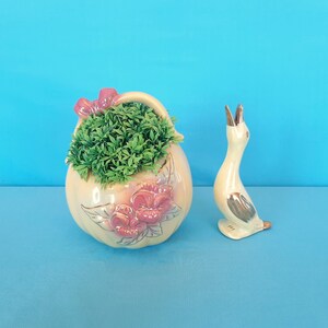 May include: A ceramic planter shaped like a pumpkin with a green plant inside and floral details. A ceramic bird figurine is next to the planter.