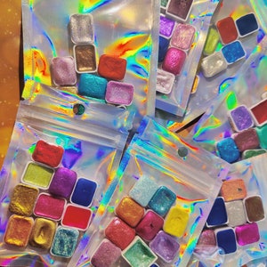 Pode incluir: Várias bolsas de plástico transparentes cheias de pequenos recipientes quadrados de tinta brilhante colorida. A tinta está em vários tons de azul, verde, vermelho, dourado e prateado.