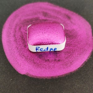 Pode incluir: Um pequeno recipiente quadrado de pó de pigmento metálico rosa brilhante com a palavra "Redne" escrita na frente.