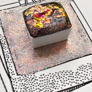 May include: A square, glittery art supply, possibly a watercolour paint pan, with a dark, textured surface and colourful iridescent flakes. It sits on a glittery, rectangular surface, clipped to a drawing with a paper clip.