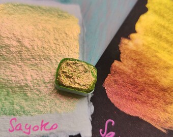 May include: Two samples of iridescent gold paint on white and black paper. The paint is a shimmery gold with a green and pink sheen. The word "Sayoko" is written in pink on both papers.