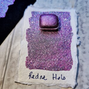 Pode incluir: Um pequeno recipiente quadrado de tinta de glitter rosa iridescente com o texto "Redne Holo" escrito em um fundo de papel branco.