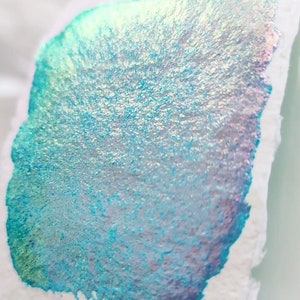 May include: A close-up of a light blue ink sample with iridescent shimmer. The ink is spread out on a white surface, showing its texture and sheen.
