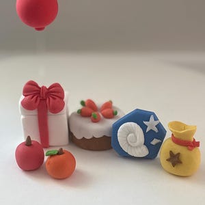 K&ouml;nnte beinhalten: Eine Sammlung von Miniatur-Tonfiguren, bestehend aus einer wei&szlig;en Geschenkbox mit roter Schleife und schwebendem roten Ballon, einem Karottenkuchen, einem roten Apfel, einer Orange, einem blauen Fossil-Symbol mit wei&szlig;er Muschel und Stern sowie einem gelben Geldsack.