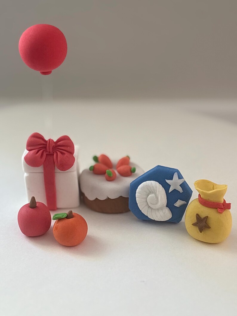 K&ouml;nnte beinhalten: Eine Sammlung von Miniatur-Tonfiguren, bestehend aus einer wei&szlig;en Geschenkbox mit roter Schleife und schwebendem roten Ballon, einem Karottenkuchen, einem roten Apfel, einer Orange, einem blauen Fossil-Symbol mit wei&szlig;er Muschel und Stern sowie einem gelben Geldsack.