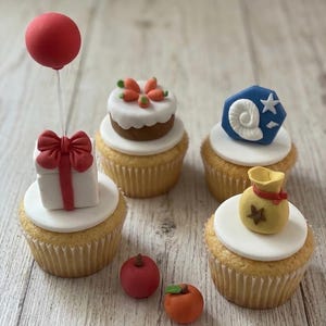 K&ouml;nnte beinhalten: Vier Cupcakes mit wei&szlig;em Fondant und thematischen Dekorationen: ein Geschenk mit rotem Ballon, ein T&ouml;rtchen mit orangefarbenen Karotten, ein blaues Sechseck mit wei&szlig;em Fossil und ein gelber Geldsack mit Stern. Zwei kleine Fr&uuml;chte liegen daneben.