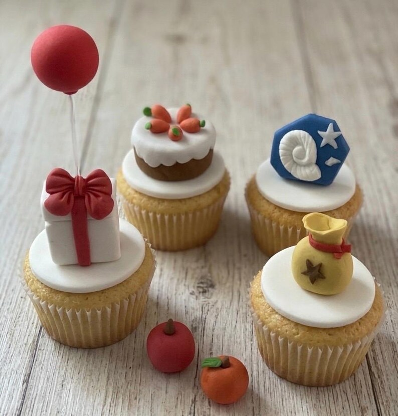K&ouml;nnte beinhalten: Vier Cupcakes mit wei&szlig;em Fondant und thematischen Dekorationen: ein Geschenk mit rotem Ballon, ein T&ouml;rtchen mit orangefarbenen Karotten, ein blaues Sechseck mit wei&szlig;em Fossil und ein gelber Geldsack mit Stern. Zwei kleine Fr&uuml;chte liegen daneben.