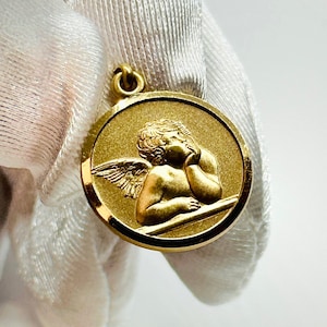 May include: A gold-colored pendant featuring a cherub with wings, resting its head on its hand. The cherub is depicted in a circular design with a raised border.