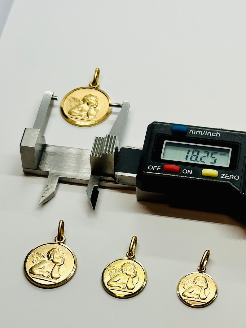 May include: Four gold angel pendants of varying sizes. The largest pendant is being measured with a digital caliper, which reads 18.25 mm.
