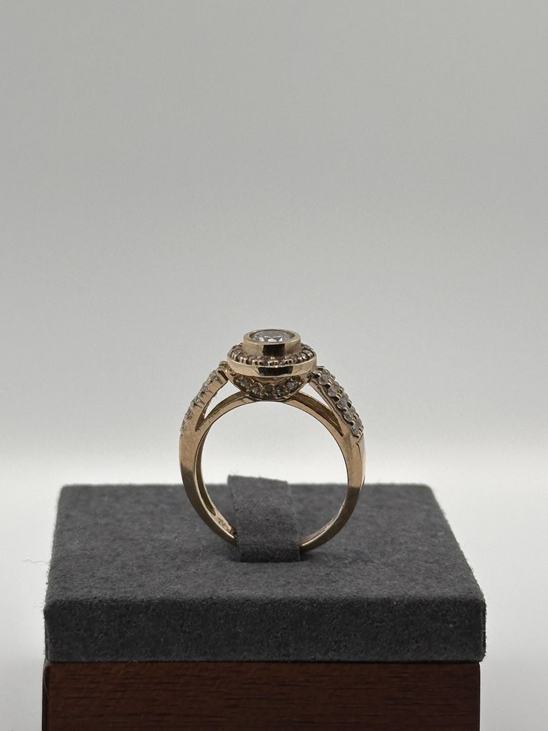May include: A gold ring with a large, clear, round gemstone surrounded by smaller gemstones. The ring is on a gray velvet ring box.