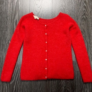 May include: A bright red long-sleeved cardigan sweater with a round neckline and a front button closure featuring seven small white buttons. The garment has a textured knit construction and a visible brand label at the inner back neckline.