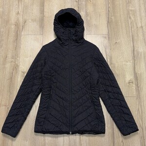 Puede incluir: Chaqueta negra acolchada con capucha. La chaqueta tiene una cremallera completa y dos bolsillos laterales. El patrón acolchado crea una apariencia texturizada. La chaqueta está colocada sobre una superficie de madera.