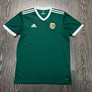 May include: A dark green short-sleeved athletic jersey with a white V-neck collar and white sleeve cuffs. The shirt features three white stripes on each shoulder, a white Adidas logo on the right chest, and a colorful team crest on the left chest.
