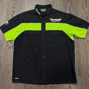 May include: A black short-sleeve button-down shirt featuring a bright lime green horizontal chest panel. The shirt displays the white text Kawasaki Racing Team on the chest, Kawasaki on the collar, and a small logo on the right sleeve and bottom hem.