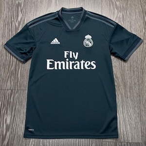 May include: A dark charcoal gray short-sleeve athletic jersey featuring the white Adidas logo on the right chest and the Real Madrid club crest on the left chest. The text Fly Emirates is printed in large white letters across the center chest.
